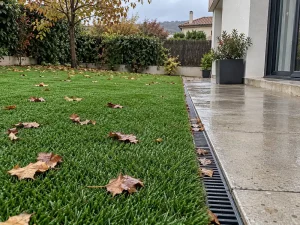 Césped artificial húmedo tras la lluvia, con hojas de otoño y un desagüe sin charcos